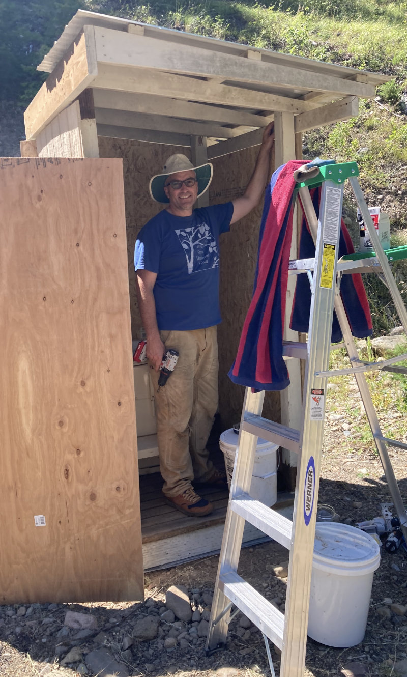 Joe, drill in hand, mid-build inside the frame of a small shed on a sunny hillside